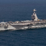 Aircraft carrier at sea with many fighter jets parked on the flight deck and a tall island superstructure.