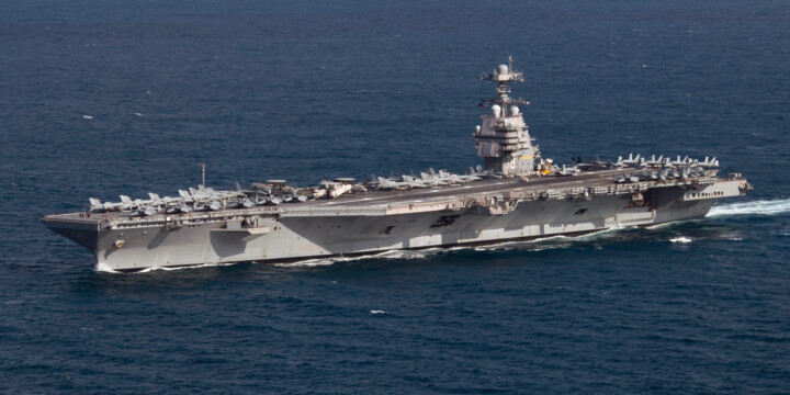 Aircraft carrier at sea with many fighter jets parked on the flight deck and a tall island superstructure.
