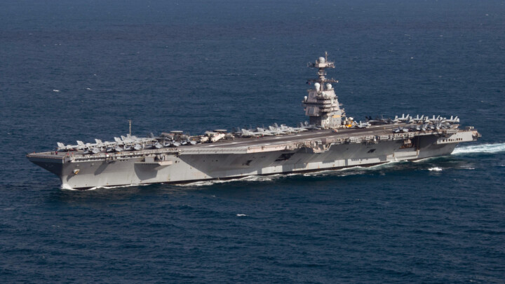 Aircraft carrier at sea with many fighter jets parked on the flight deck and a tall island superstructure.